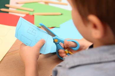 Photo of Little boy cutting color paper with scissors at table indoors, closeup
