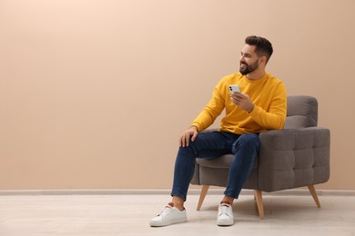 Photo of Handsome man with smartphone sitting in armchair near beige wall indoors, space for text
