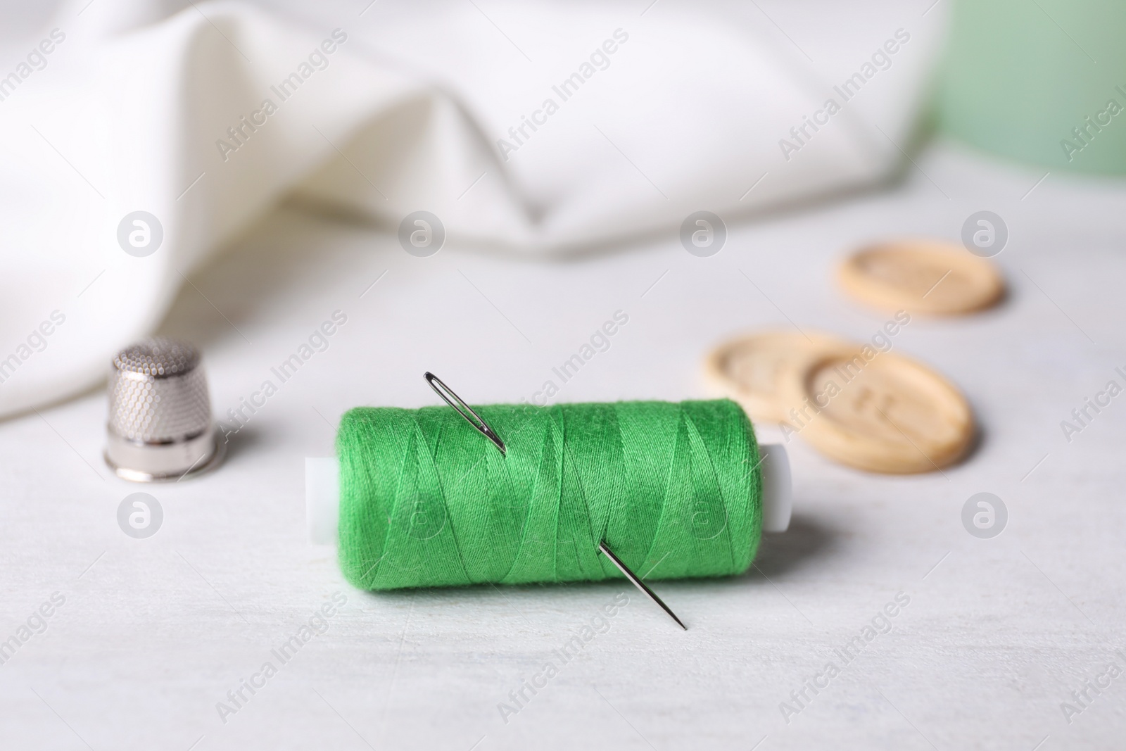 Photo of Spool of green sewing thread with needle, thimble and buttons on white table