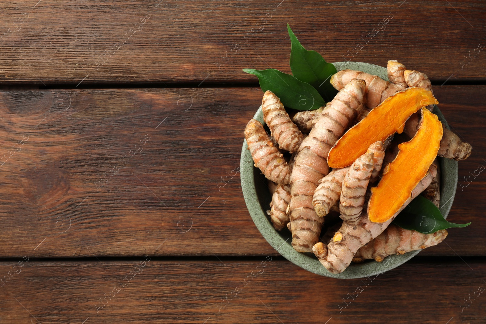 Photo of Bowl with fresh turmeric roots and leaves on wooden table, top view. Space for text