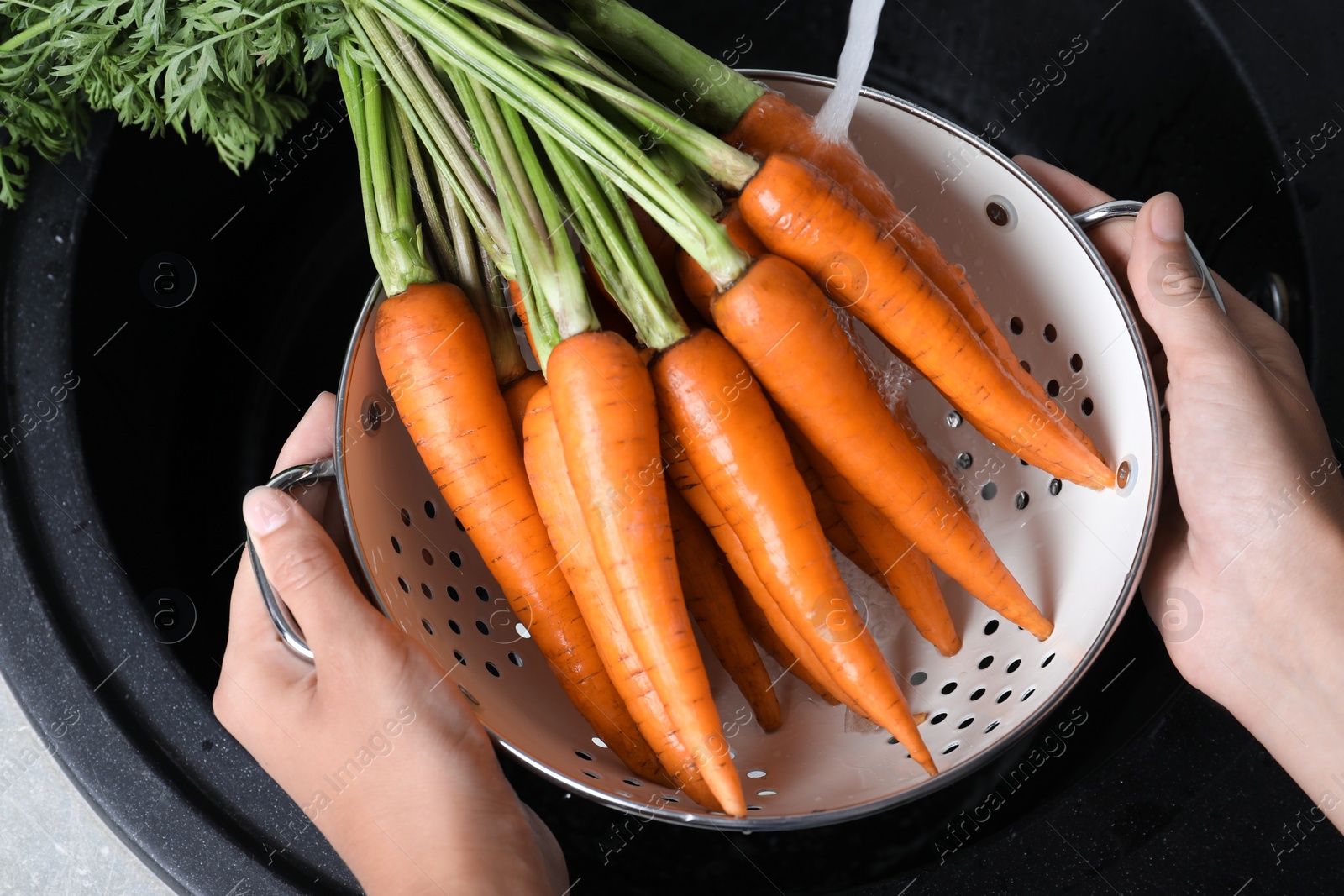 Photo of Woman washing ripe carrots in colander with running water over sink, closeup