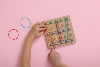 Photo of Motor skills development. Boy playing with geoboard and rubber bands at pink table, top view