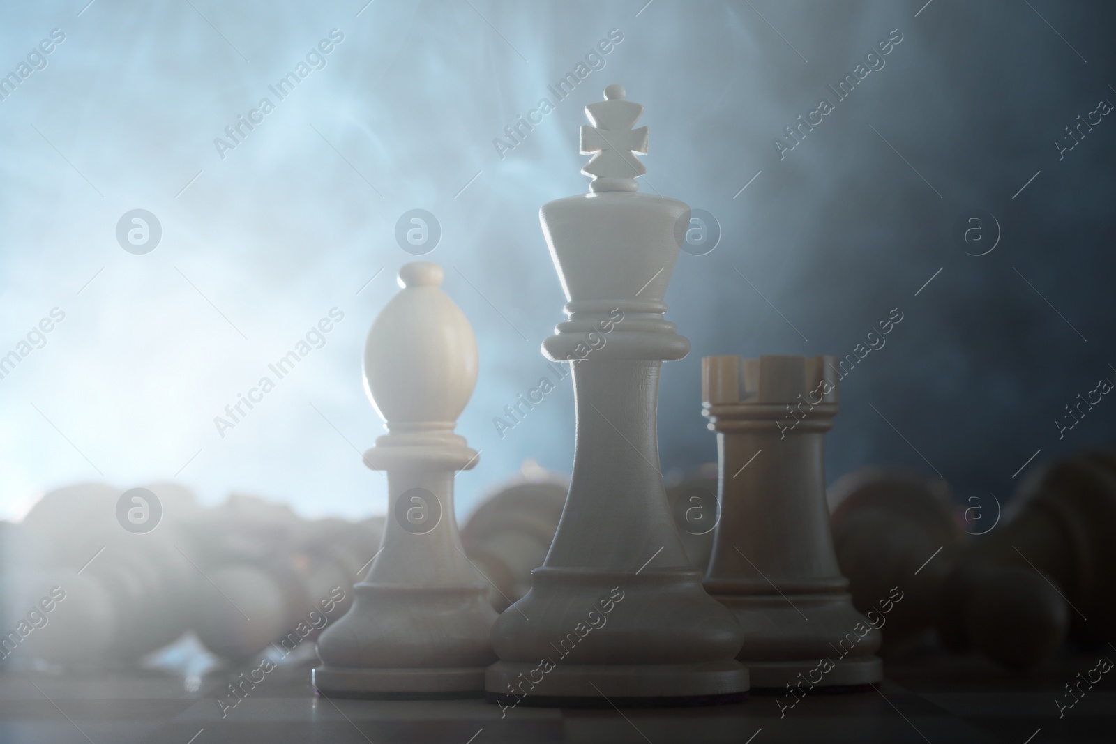 Image of Wooden king, rook and bishop among fallen chess pieces on checkerboard in smoke