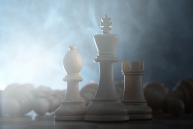 Wooden king, rook and bishop among fallen chess pieces on checkerboard in smoke