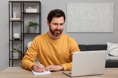 Man using laptop and writing something in notebook at wooden table indoors