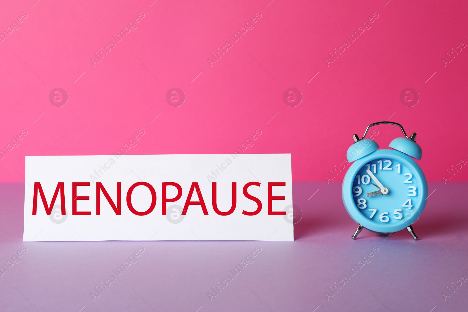 Photo of Card with word Menopause and alarm clock on violet table against pink background