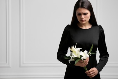 Sad woman with lilies mourning near white wall, space for text. Funeral ceremony