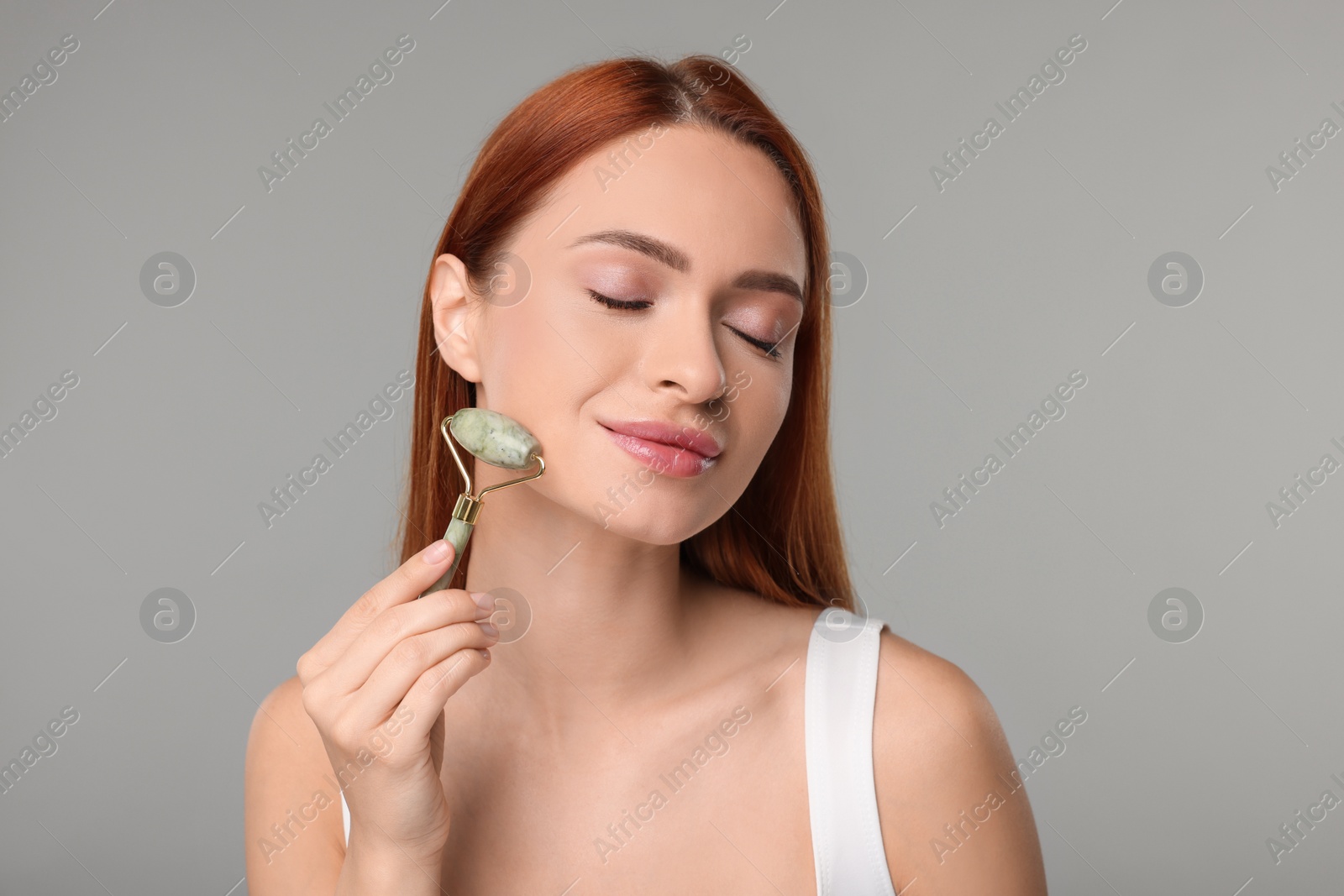 Photo of Young woman massaging her face with jade roller on grey background