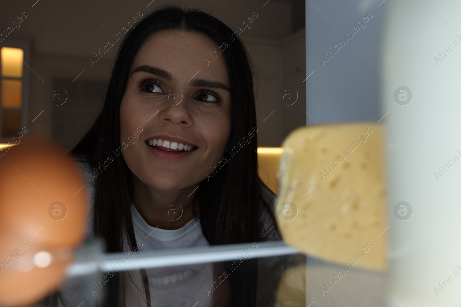 Photo of Young woman near modern refrigerator in kitchen at night, view from inside