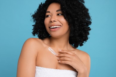 Young woman applying cream onto body on light blue background