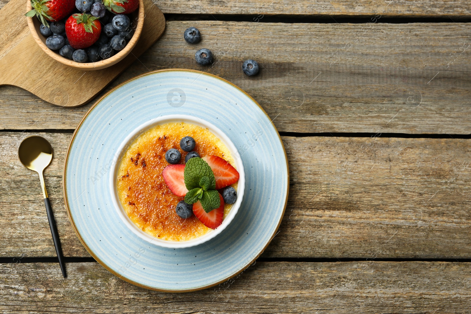 Photo of Delicious creme brulee with berries and mint in bowl served on wooden table, flat lay. Space for text