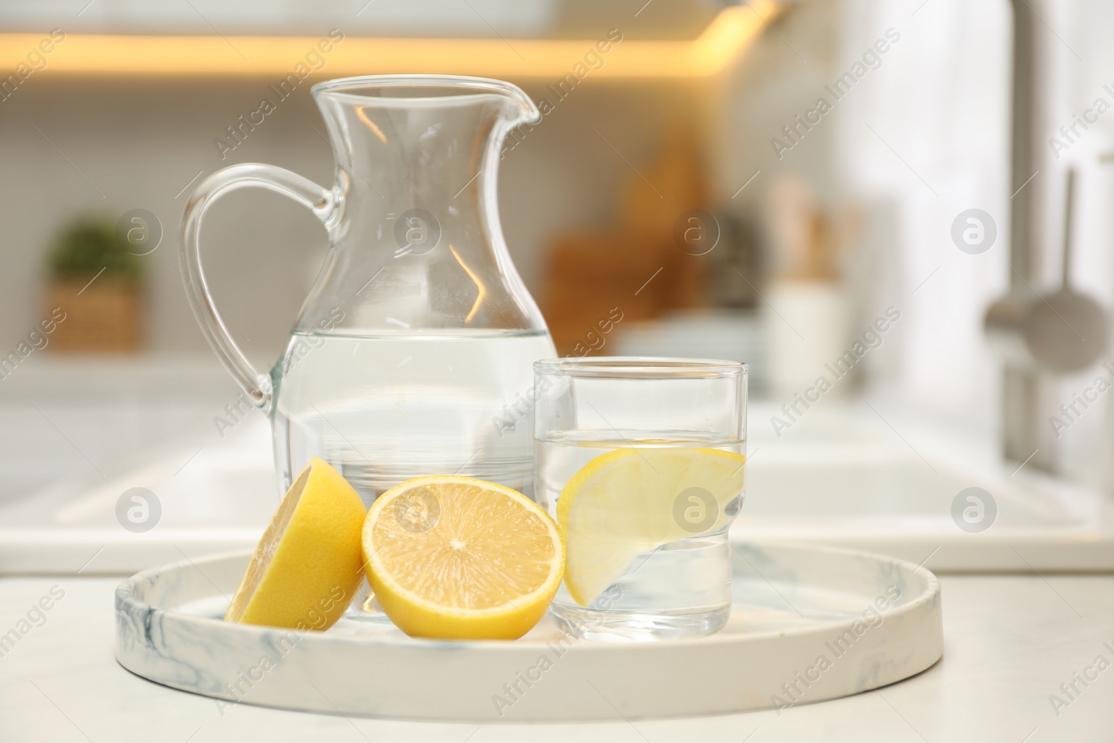 Photo of Jug, glass with clear water and lemons on white table in kitchen