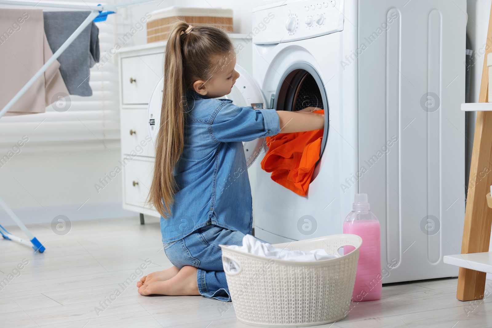 Photo of Little girl putting dirty clothes into washing machine in bathroom