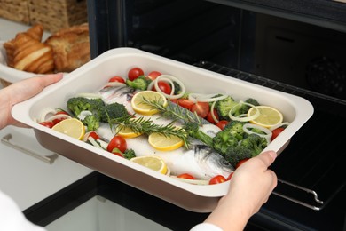 Photo of Woman putting baking dish with raw fish and vegetables into oven in kitchen, closeup