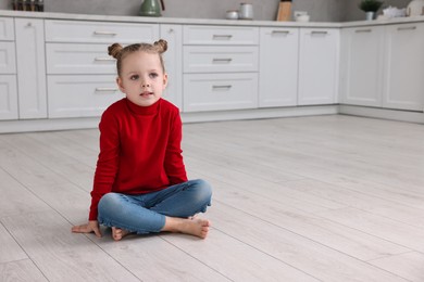 Photo of Cute little girl sitting on warm floor in kitchen, space for text. Heating system