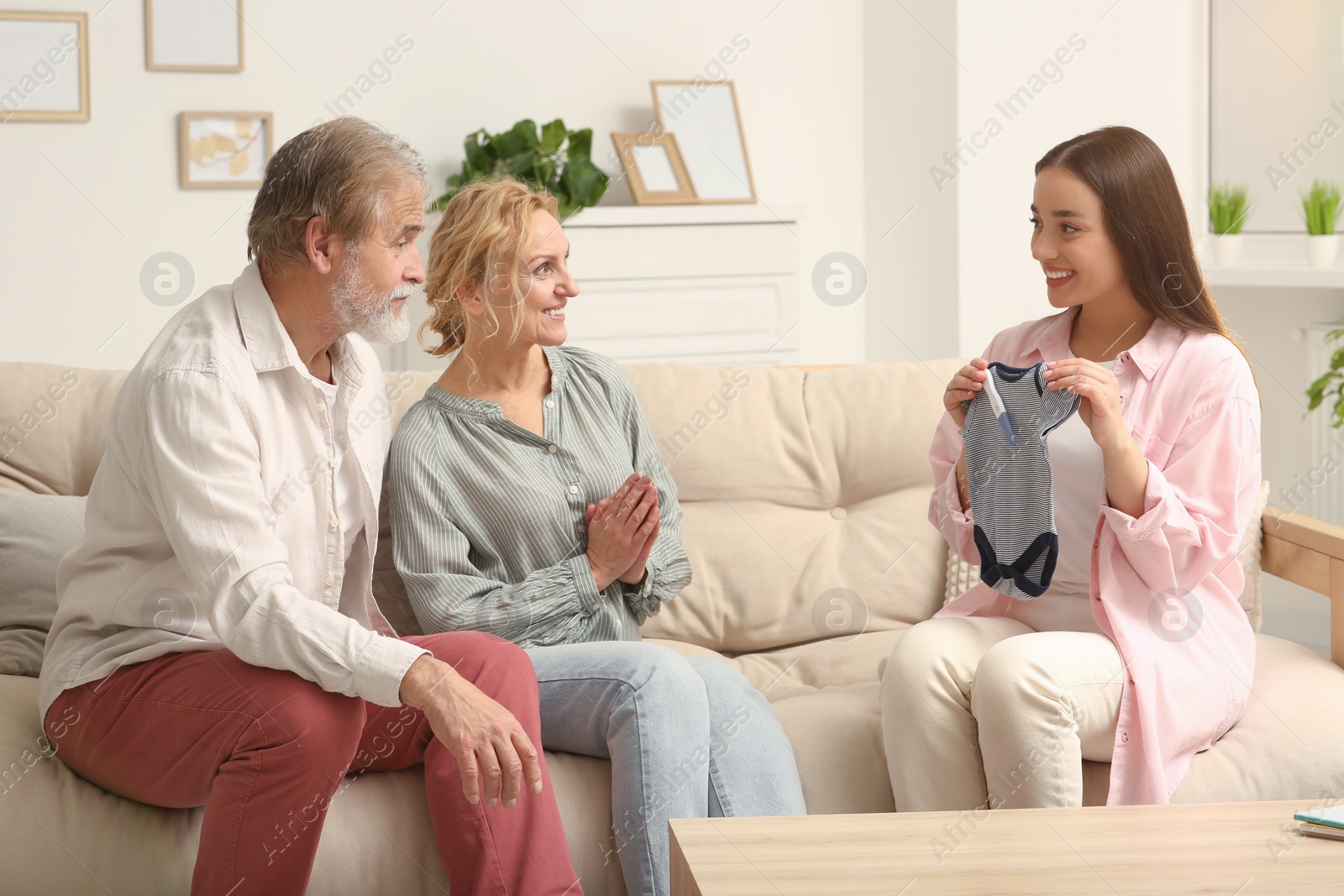 Photo of Happy woman showing her parents baby's bodysuit at home. Grandparents' reaction to future grandson