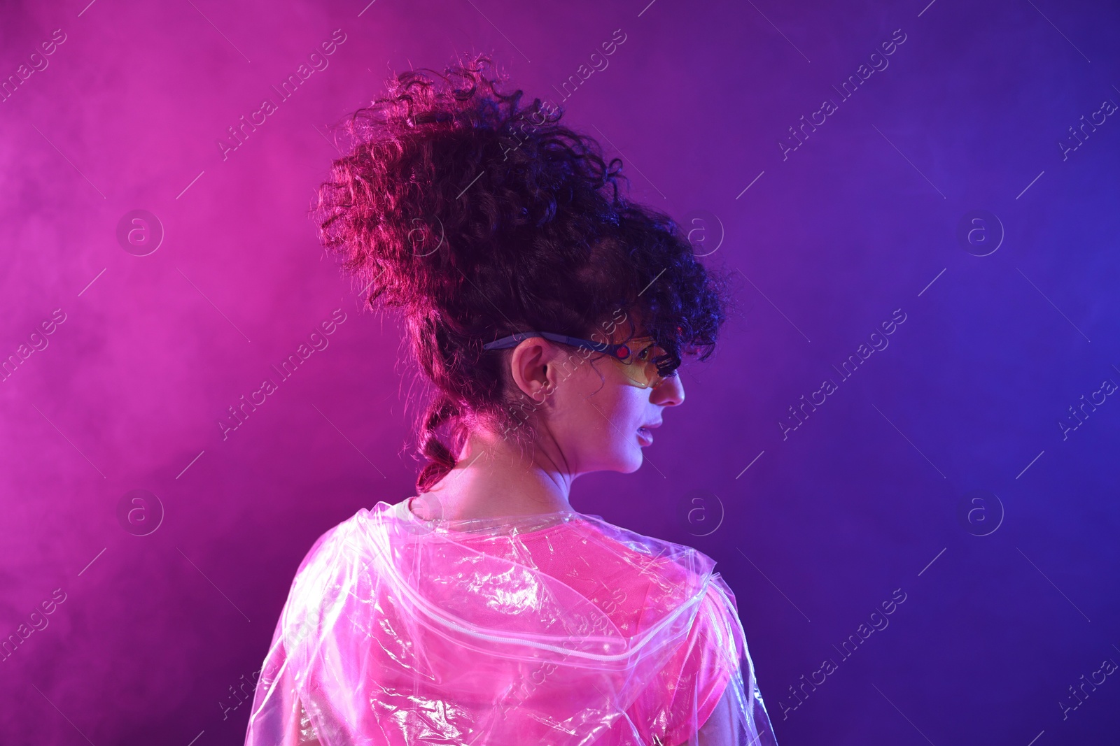 Photo of Beautiful young woman in transparent coat and sunglasses posing on color background in neon lights and smoke