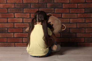Photo of Child abuse. Upset little girl with teddy bear sitting on floor near brick wall indoors, back view