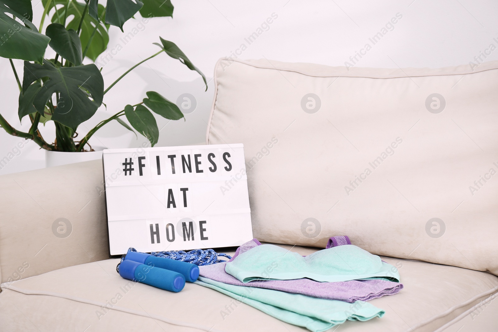 Photo of Sport equipment and lightbox with hashtag FITNESS AT HOME on sofa indoors. Message to promote self-isolation during COVID‑19 pandemic