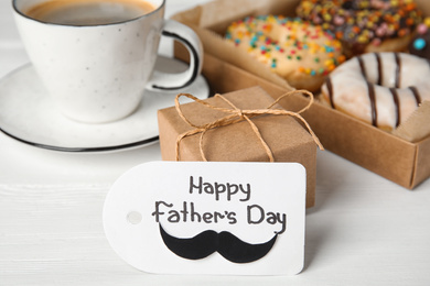 Photo of Tag with phrase HAPPY FATHER'S DAY, donuts, coffee and gift on white wooden table