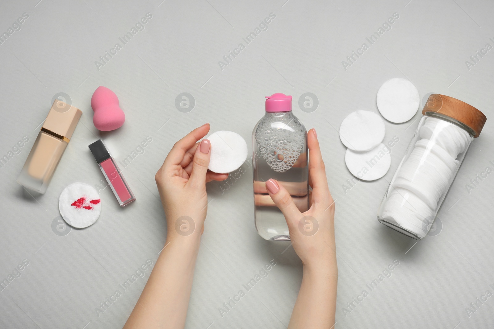 Photo of Woman with makeup remover, cotton pads, foundation, lip gloss and sponge on light grey background, top view