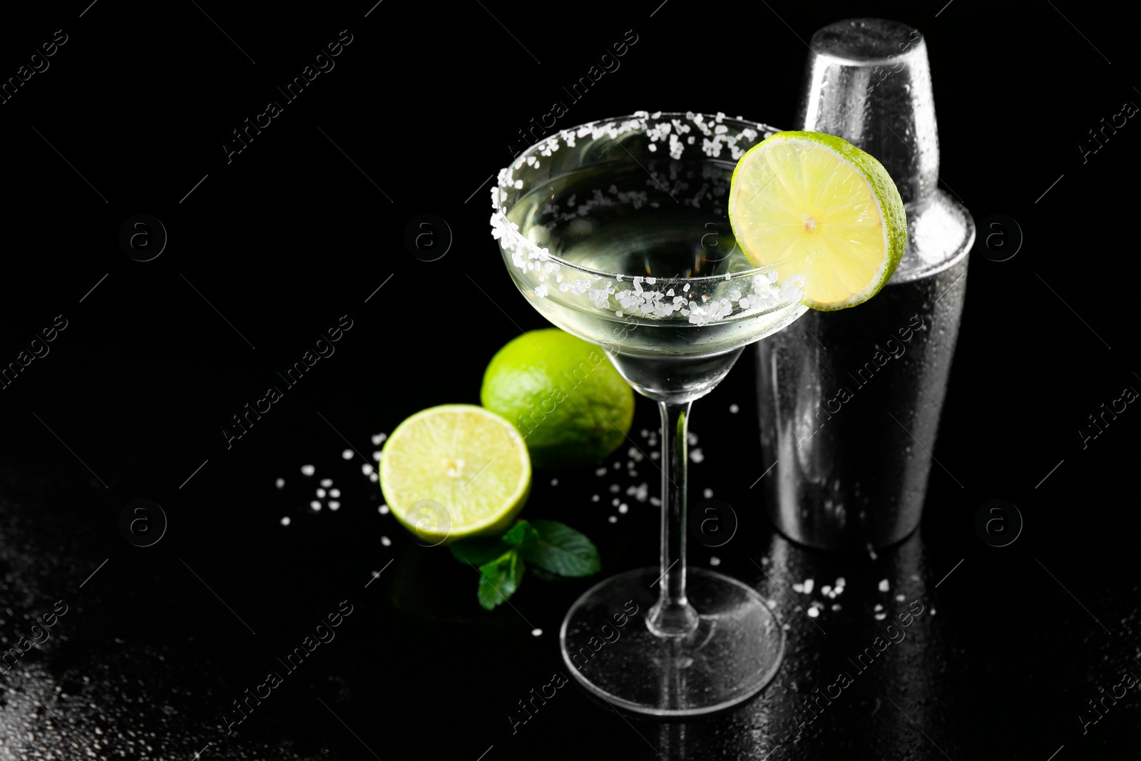 Photo of Metal shaker, delicious cocktail, limes and mint on black mirror surface, space for text