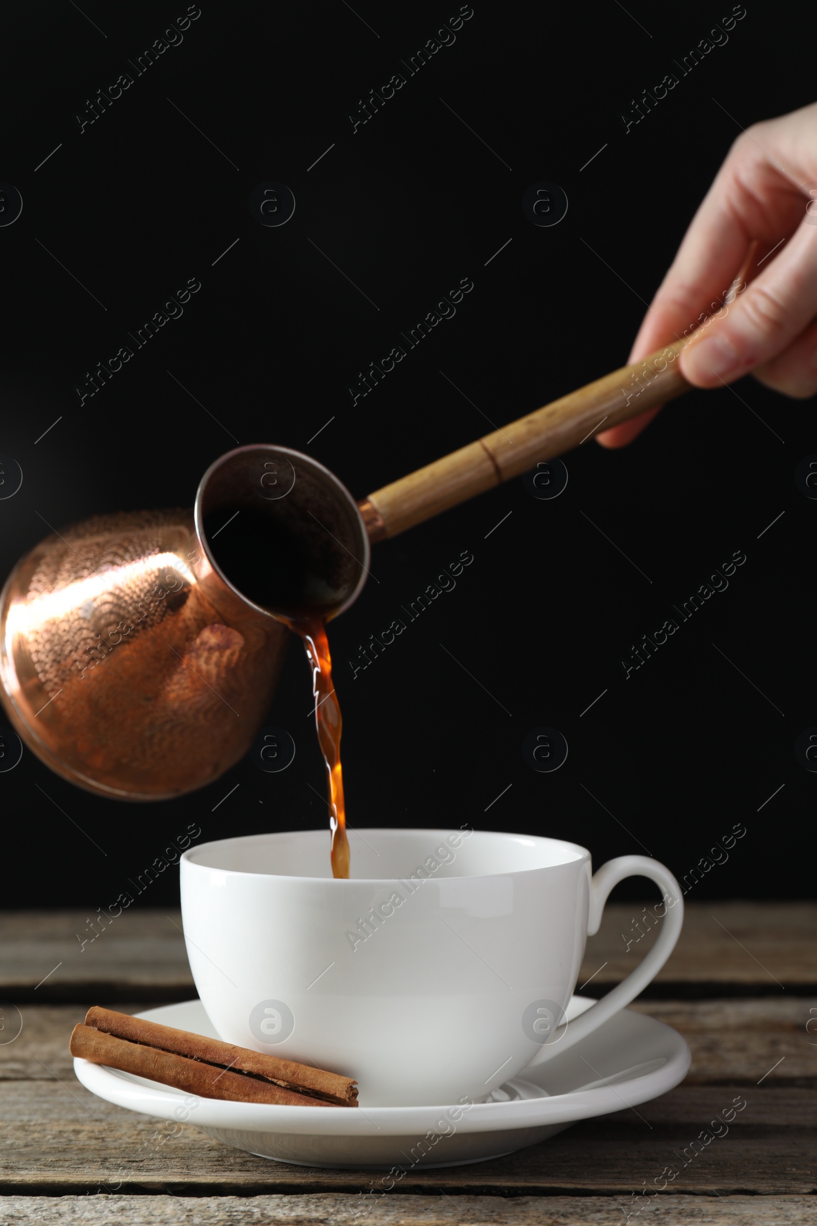 Photo of Turkish coffee. Woman pouring brewed beverage from cezve into cup at wooden table against black background, closeup