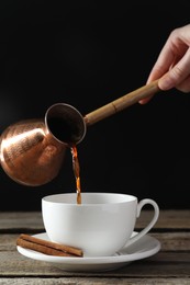 Turkish coffee. Woman pouring brewed beverage from cezve into cup at wooden table against black background, closeup