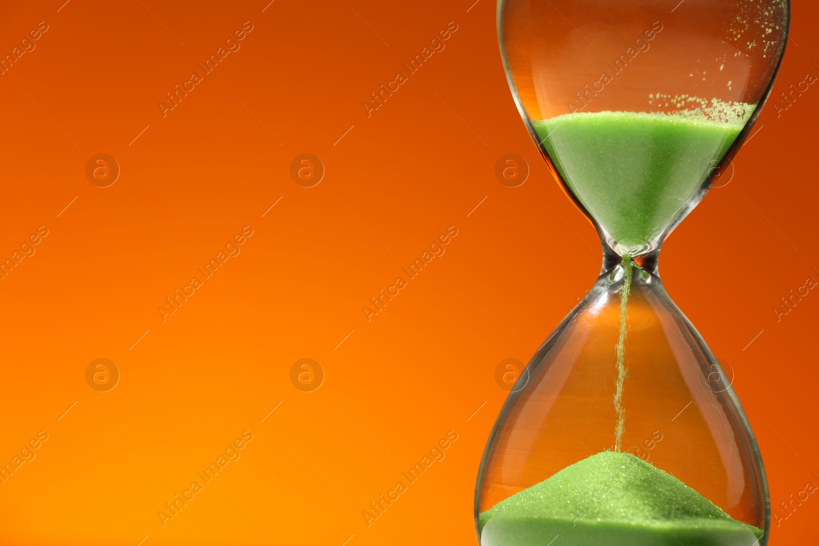 Photo of Hourglass with light green flowing sand against orange background, closeup. Space for text
