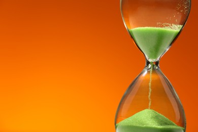 Hourglass with light green flowing sand against orange background, closeup. Space for text