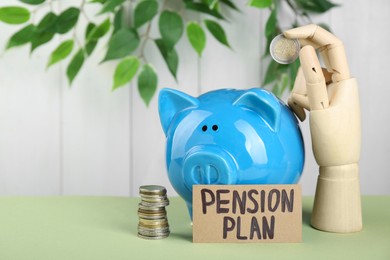Photo of Card with phrase Pension Plan, wooden mannequin hand holding coin and piggy bank on light green table. Retirement concept