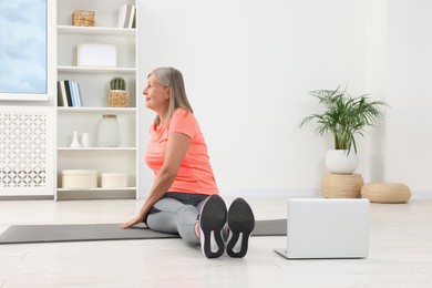 Senior woman in sportswear stretching near laptop on mat at home