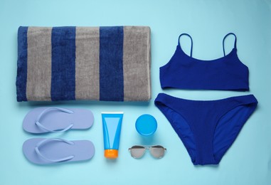 Beach towel, swimsuit, flip flops, sunglasses and sun protection product on light blue background, flat lay