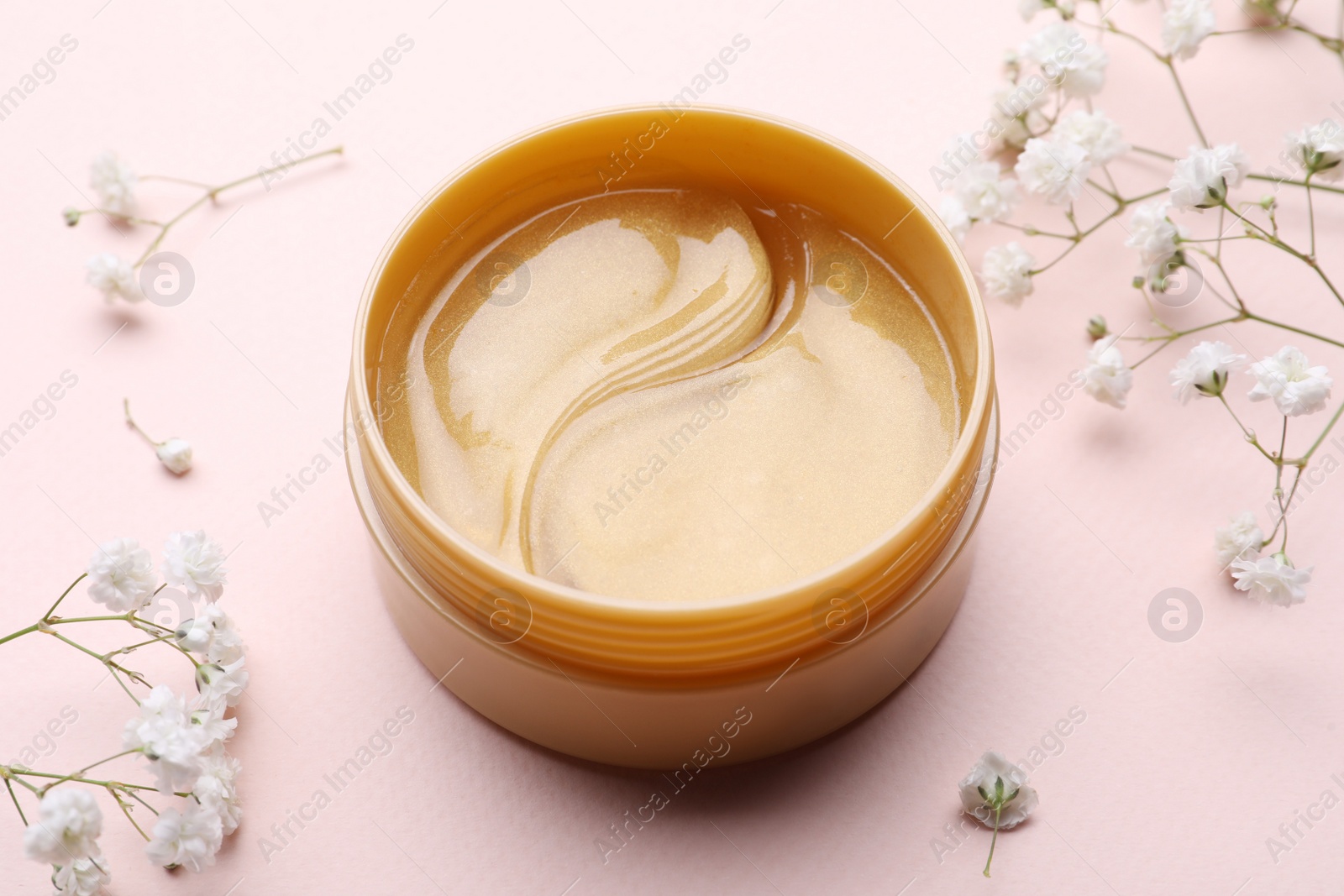 Photo of Under eye patches in jar and flowers on light pink background, closeup. Cosmetic product