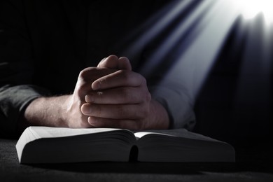 Image of Christian man praying over Bible in darkness, closeup. Holy light shining at him