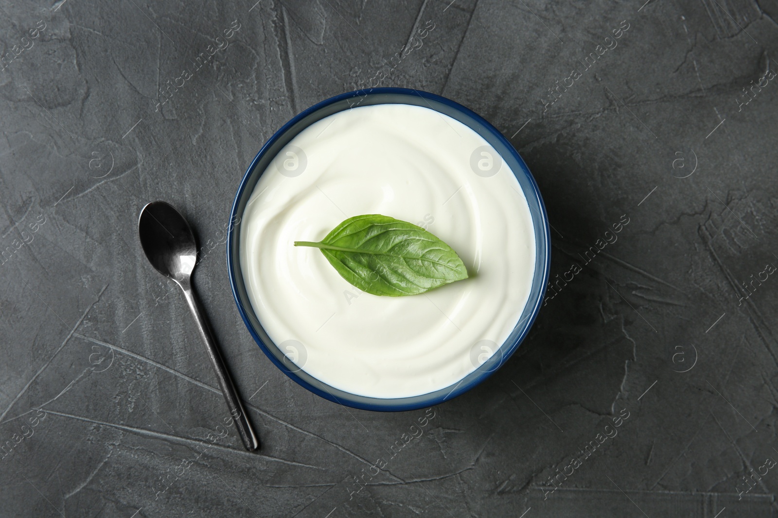 Photo of Bowl of fresh sour cream with basil and spoon on grey table, top view