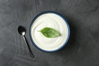 Bowl of fresh sour cream with basil and spoon on grey table, top view