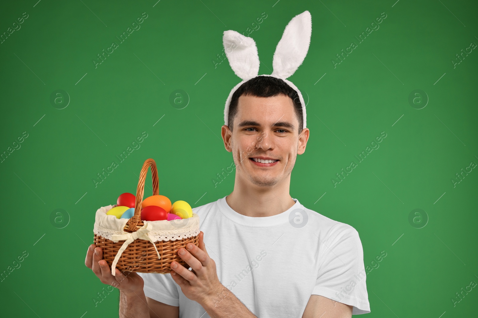 Photo of Easter celebration. Handsome young man with bunny ears holding basket of painted eggs on green background