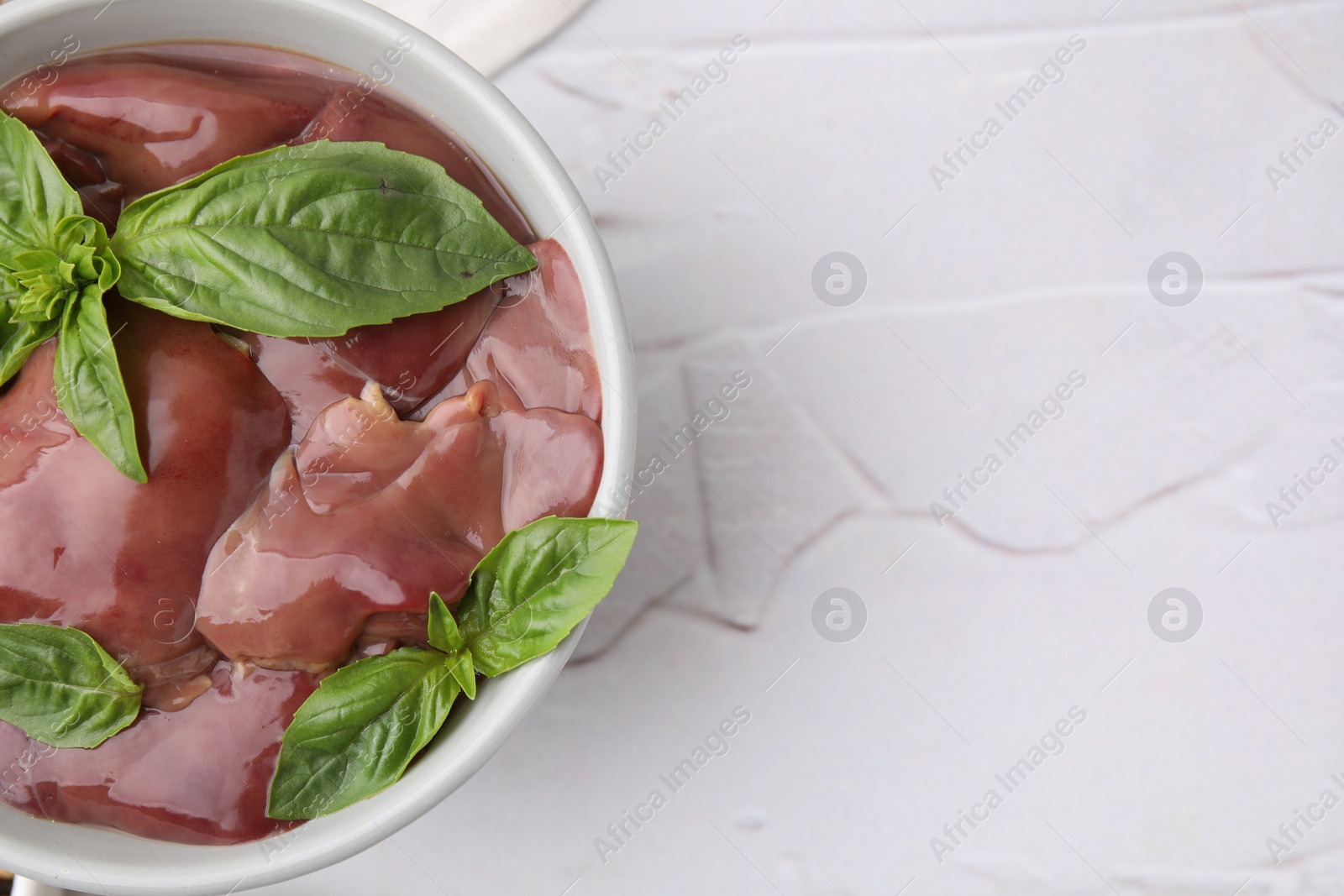 Photo of Bowl with raw chicken liver and basil on white textured table, top view. Space for text