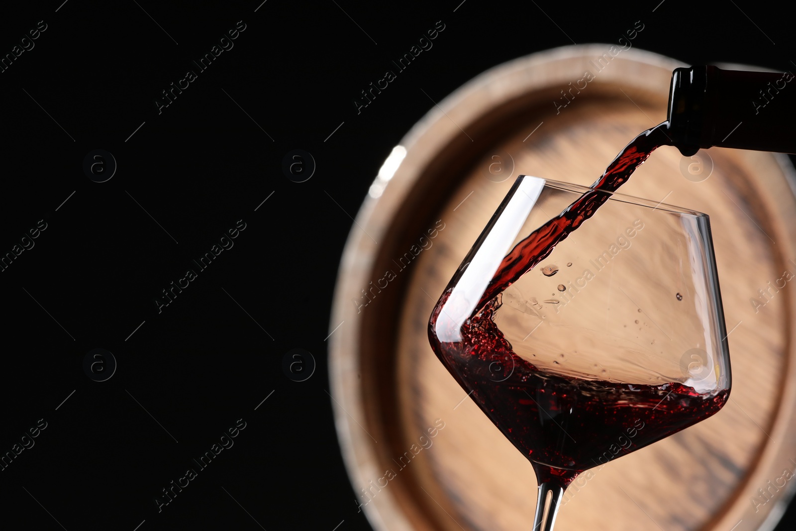 Photo of Pouring red wine from bottle into glass near wooden barrel against black background, closeup. Space for text