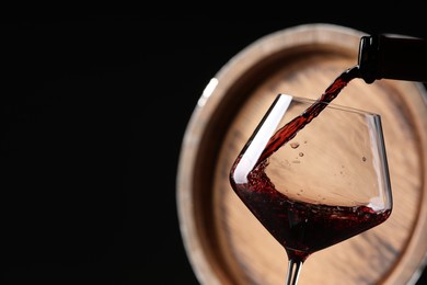 Pouring red wine from bottle into glass near wooden barrel against black background, closeup. Space for text