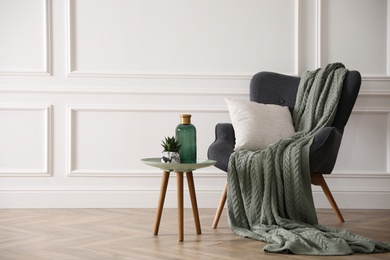 Modern armchair with knitted blanket and cushion in stylish room interior. Space for text