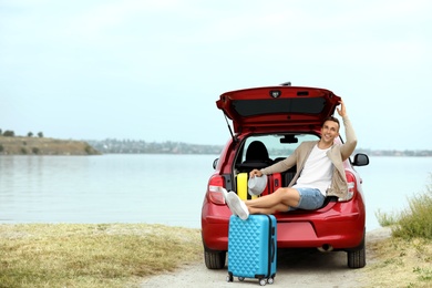 Photo of Happy man sitting in car trunk with suitcases on riverside. Space for text