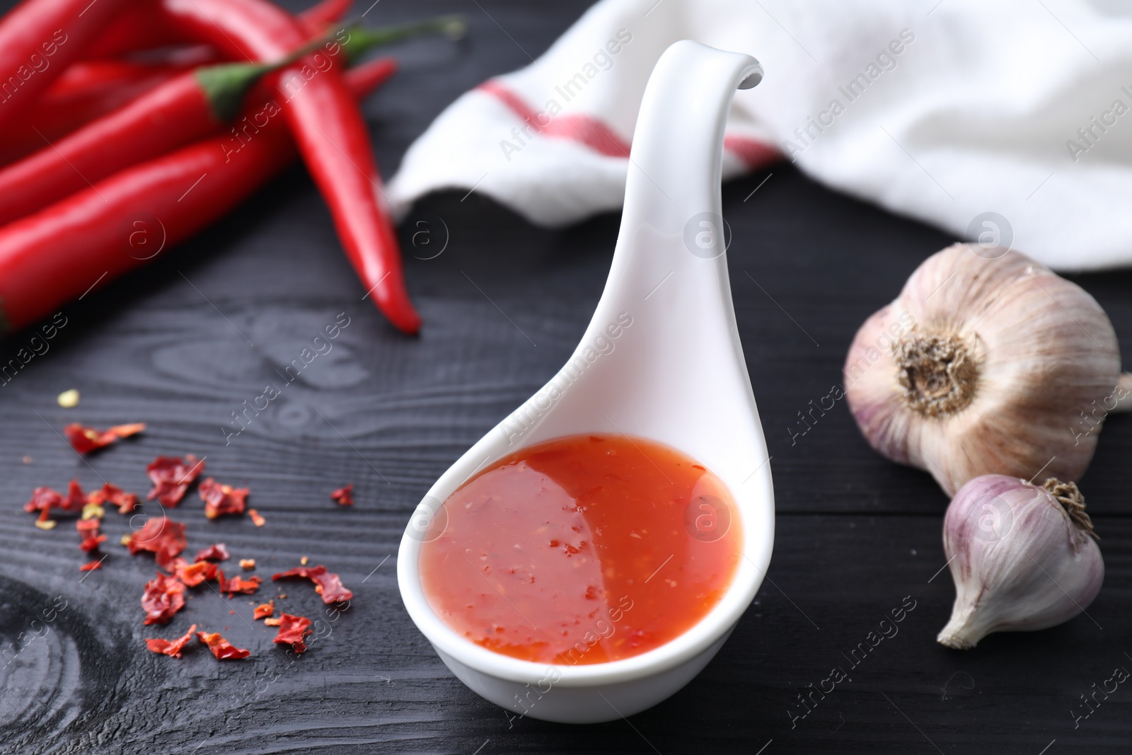 Photo of Spicy chili sauce in spoon, peppers and garlic on black wooden table, closeup