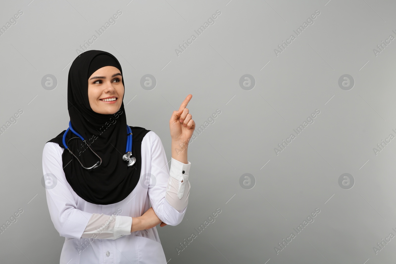 Photo of Muslim woman in hijab and medical uniform pointing at something on light gray background, space for text