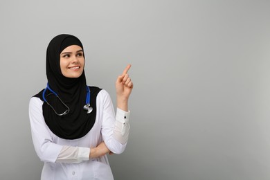 Muslim woman in hijab and medical uniform pointing at something on light gray background, space for text