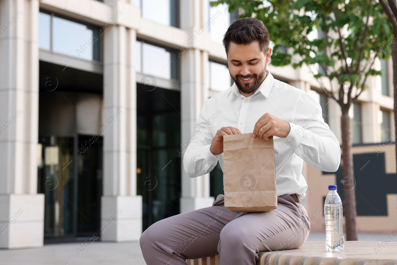 Photo of Happy businessman with paper bag having lunch on bench outdoors