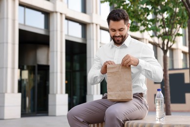 Happy businessman with paper bag having lunch on bench outdoors