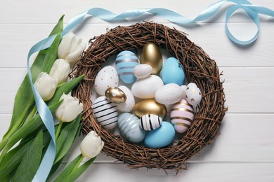 Many painted Easter eggs, tulip flowers and ribbon on white wooden table, flat lay