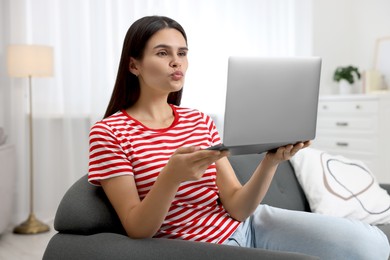 Young woman having video chat via laptop and blowing kiss on sofa in room